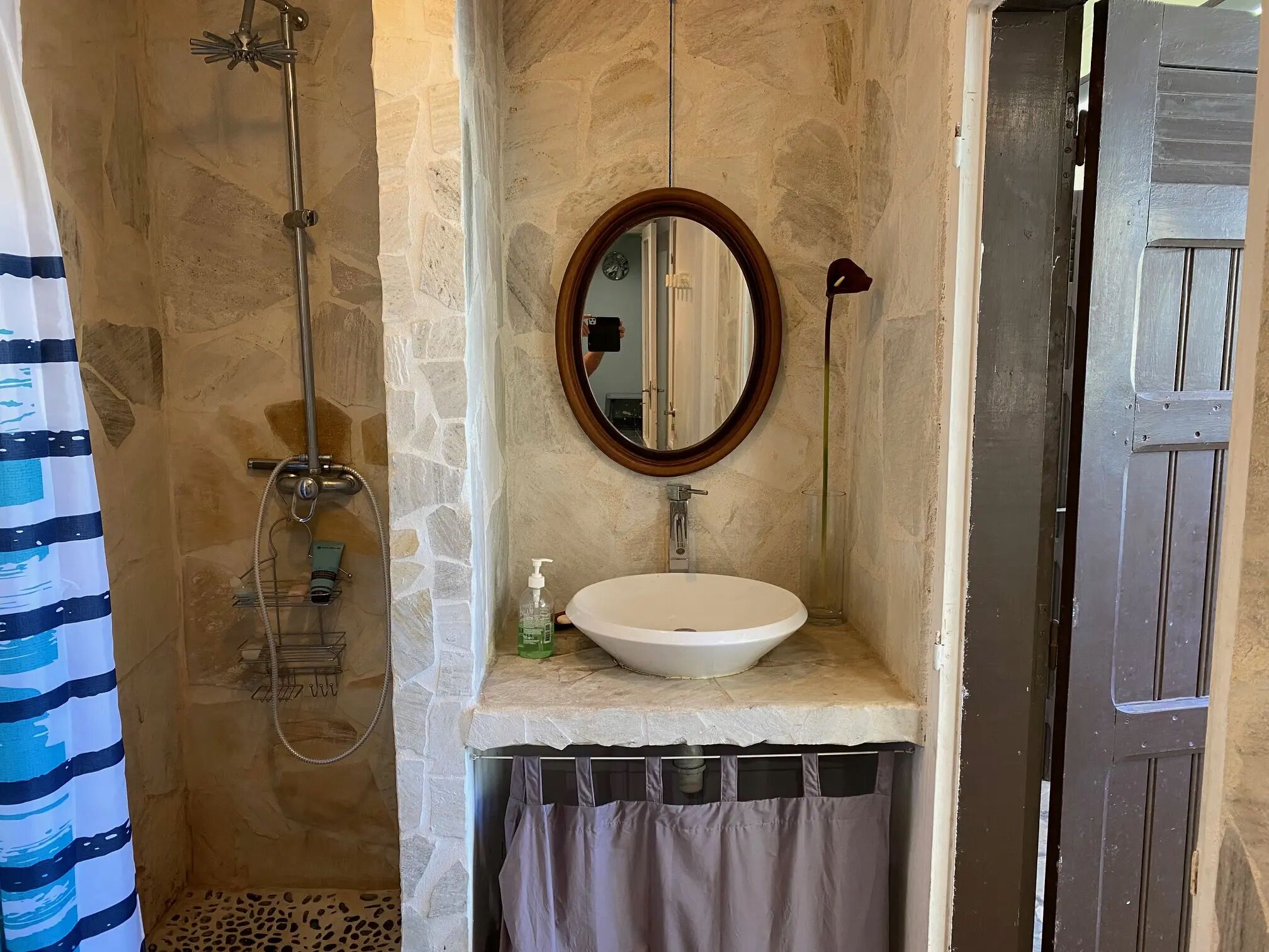 A bathroom with a stone wall, a round mirror, a white sink, and a shower.