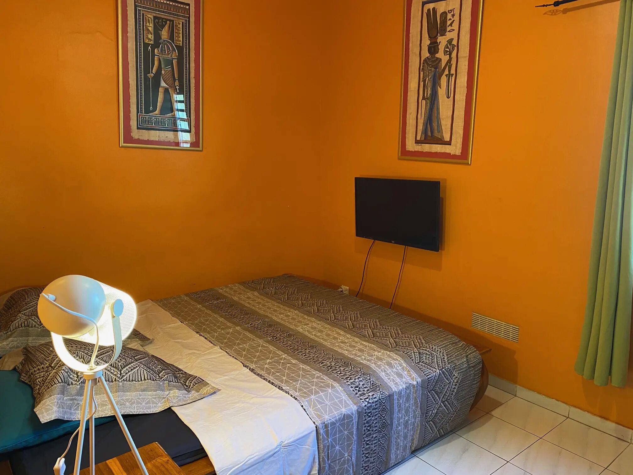Une pièce avec un mur orange, un lit recouvert d’une literie à motifs, un téléviseur fixé au mur et une petite table avec une lampe.