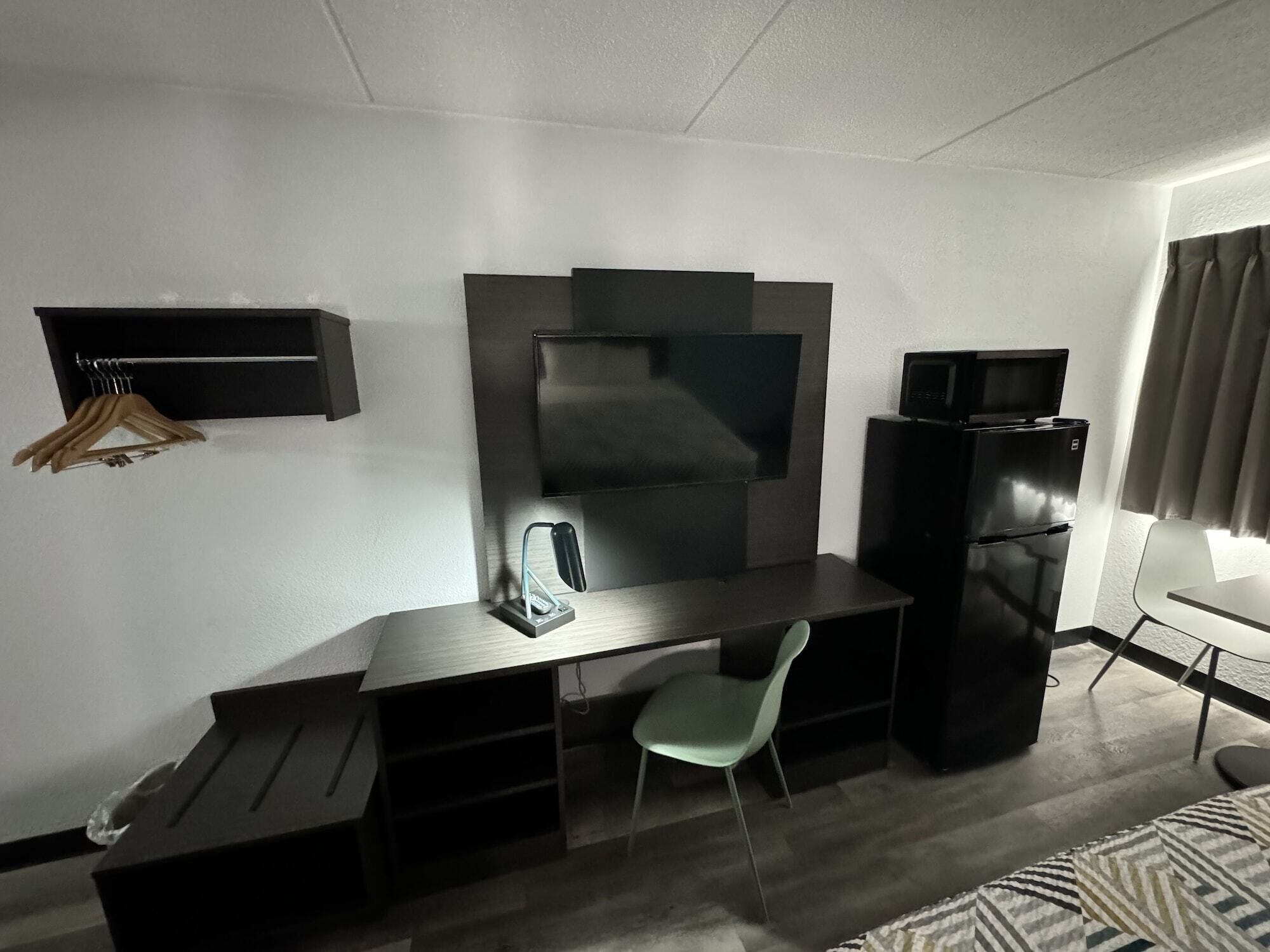 A hotel room with a flat-screen TV mounted on the wall, a small refrigerator, a desk with a chair, and a bed with patterned bedding.