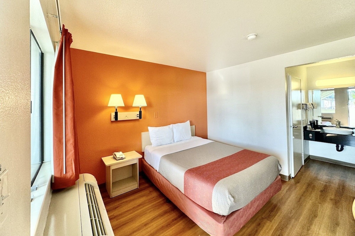 A hotel room with a bed, bedside table, lamp, and a bathroom with a sink and mirror.