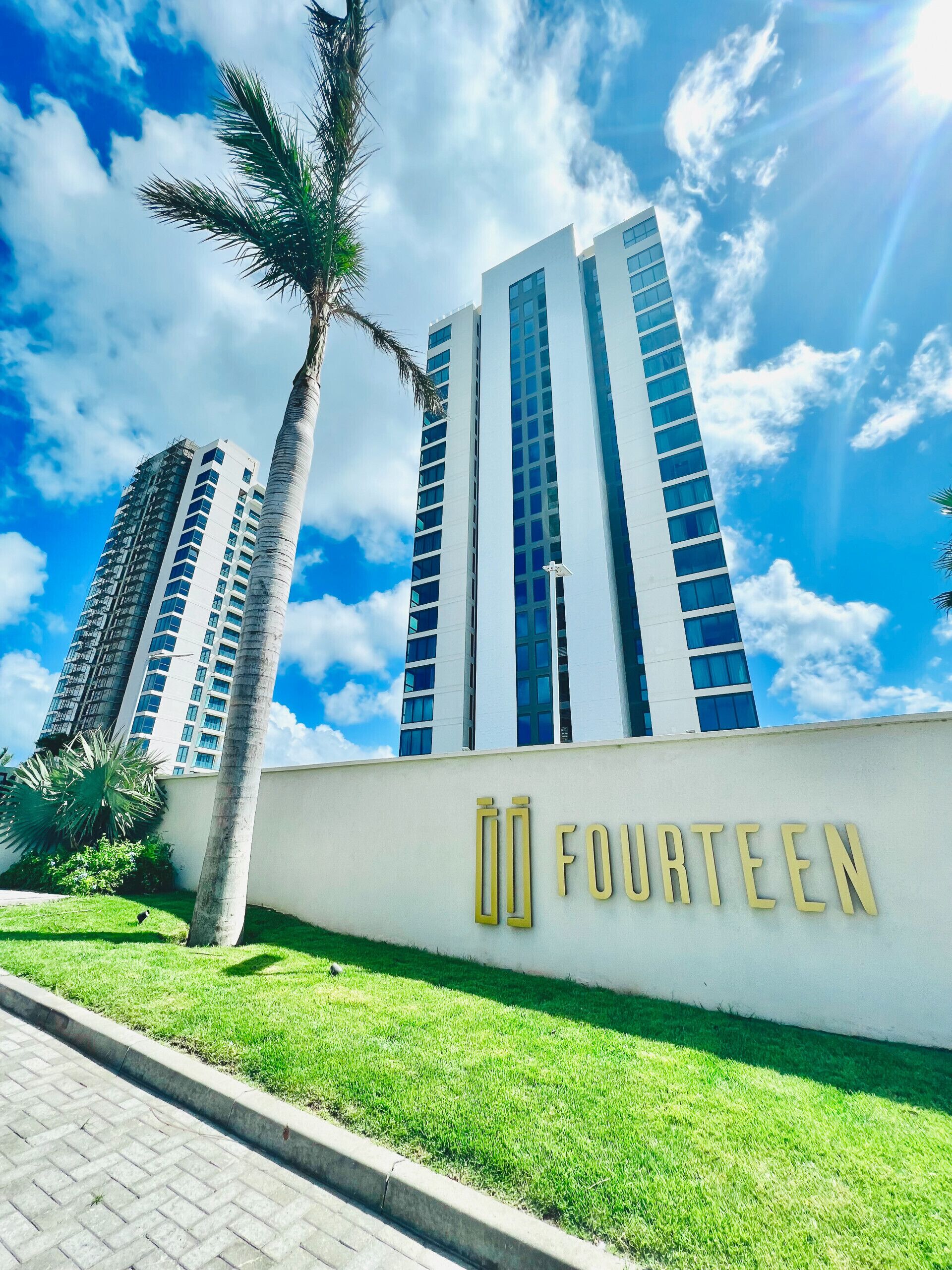 Un edificio moderno con un letrero que dice “FOURTEEN” en letras doradas, rodeado de palmeras y un cielo azul despejado.
