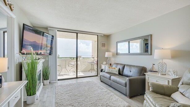 A modern living room with a grey sofa, a white coffee table, and a TV mounted on the wall. There is a sliding glass door leading to an outdoor balcony with a table and chairs.