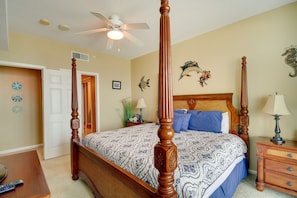 A four-poster bed with a wooden headboard, a ceiling fan, a bedside table with a lamp, and decorative wall art. - Beachfront Gulfport Vacation Rental w/ Balcony! (Gulfport)