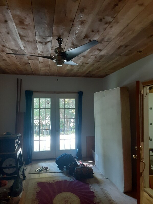 A room with wooden ceiling beams, a ceiling fan, a window with curtains, and a bed with a red and white patterned bedspread.