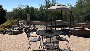 Un patio al aire libre con una mesa redonda y sillas, un área de estar cubierta y un pozo de fuego de piedra.