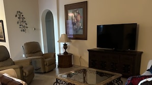 Una sala de estar con sofá, sillón, mesa de centro de vidrio, televisor y mueble.