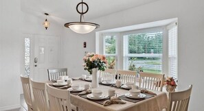 A dining room with a table set for six, white chairs, a vase with flowers, and a window with blinds.