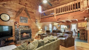 Ein gemütliches Blockhaus-Interieur mit Kamin aus Stein, Ledersofas, einem Fernseher und einem Schlafzimmer im oberen Bereich.