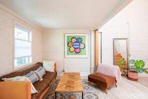A cozy living room with a brown sofa, a wooden coffee table, and a brown leather chair. There's a colorful abstract painting on the wall and a staircase leading to the upper floor.