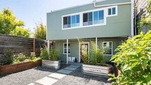 A modern two-story house with a light green exterior and white trim. There are two large windows on the second floor and a small front porch with a door. The front yard has a gravel pathway and raised garden beds with various plants. - Guesthouse, Urban Oasis, 2b/2bth (Berkeley)