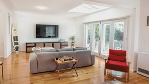 A living room with a sofa, a red chair, a wooden coffee table with a chessboard, a surfboard, and a TV on the wall.