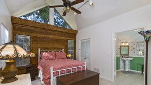 Una camera da letto con soffitto in legno, un letto con copripiumino rosa, un comodino con una lampada e vista sul bagno attraverso una porta aperta.