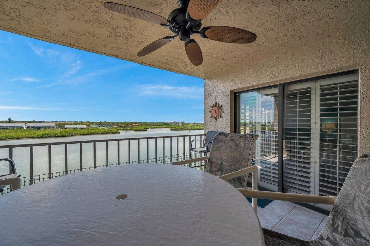 Un balcon avec un ventilateur de plafond, une table et des chaises. On peut voir une grande fenêtre avec des stores.