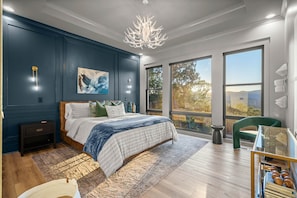 A modern bedroom with a large bed, a chandelier, a painting, and a view of the outdoors.
