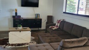 A living room with a sectional sofa, a flat-screen TV, a wooden coffee table, and a window with blinds.