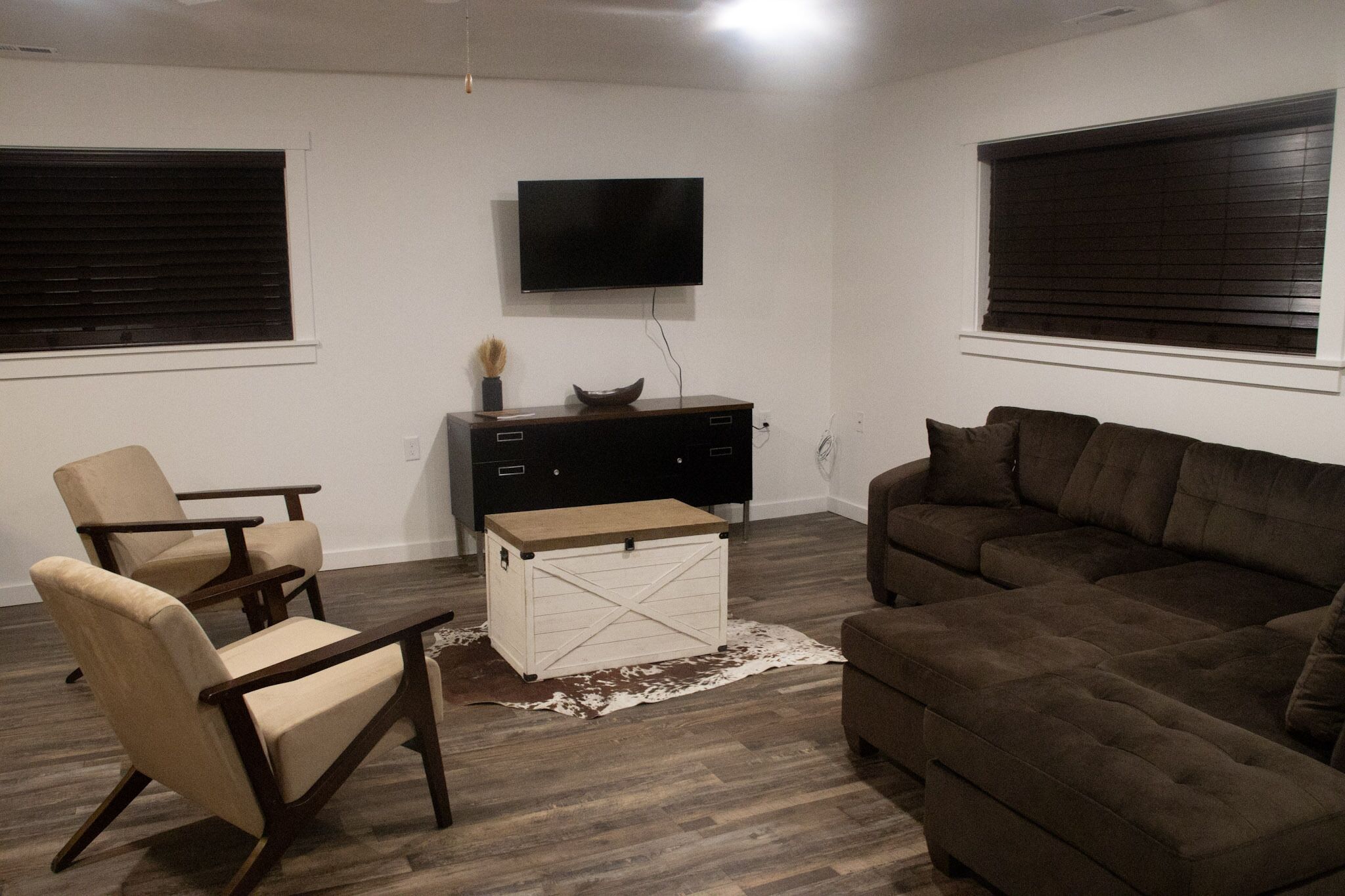 Een woonkamer met een bruine bank, twee fauteuils, een houten salontafel, een witte dressoir en een flatscreen televisie die aan de muur is bevestigd.