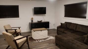 Een woonkamer met een bruine bank, twee fauteuils, een houten salontafel, een witte dressoir en een flatscreen televisie die aan de muur is bevestigd.