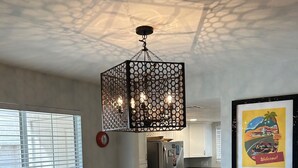 A dining area with a glass table surrounded by wicker chairs, a decorative hanging light fixture, and a kitchen in the background.