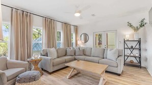 A spacious living room with a large sectional sofa, a coffee table, and a side table. There is a ceiling fan, a wall-mounted mirror, and a black shelving unit with decorative items. - Changes in Attitude | 6-Seater Golf Cart | 4 Bikes (Santa Rosa Beach)