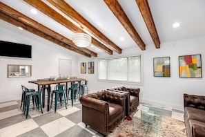 A spacious room with exposed wooden beams, a dining area with a table and chairs, and a large window with blinds.
