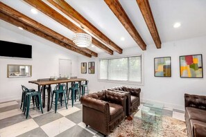 A spacious living area with a high ceiling, exposed wooden beams, a dining table with chairs, and a large brown leather sofa.