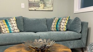 Ein Wohnzimmer mit grauem Sofa, gemusterten Kissen, einem runden Couchtisch mit Metallschale und einem Blumenbild an der Wand.