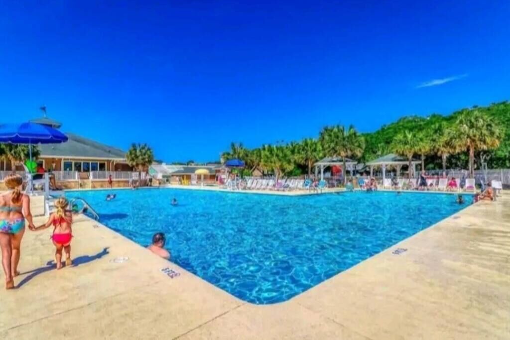 Ein Swimmingpool mit Liegestühlen und Sonnenschirmen, umgeben von Palmen und einem wolkenlosen, blauen Himmel.