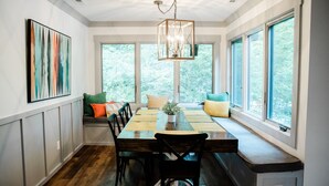 Un comedor con una mesa de madera, sillas y un banco con cojines. Amplias ventanas ofrecen vistas a árboles.