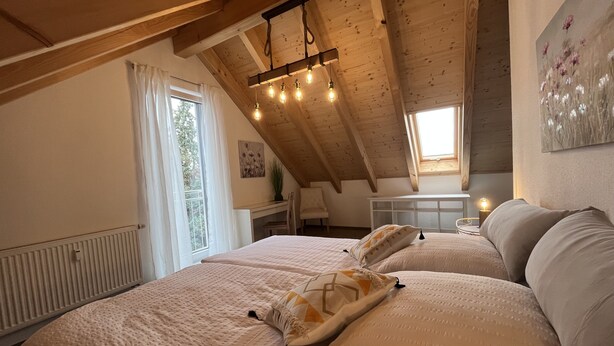 A cozy attic bedroom with a sloped ceiling, a large bed with a quilted cover, a desk with a chair, and a window with curtains. - Wohnung Carmen (Bad Rappenau)