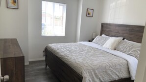 Une chambre à coucher avec un grand lit, une tête de lit en bois, une fenêtre avec des stores et une petite table de chevet avec une lampe.