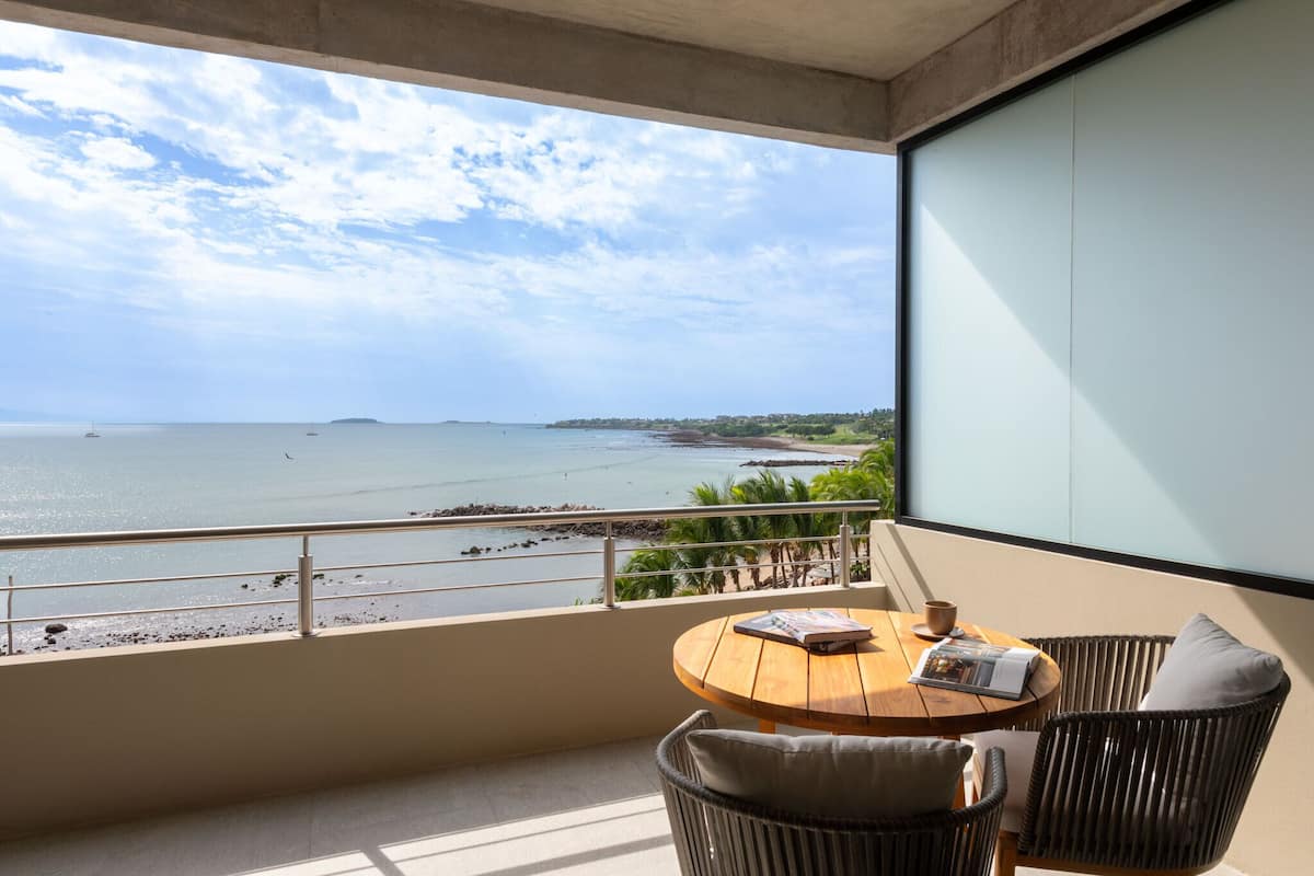 Un balcón con vista al mar, una mesa de madera redonda, sillas de mimbre y una revista.