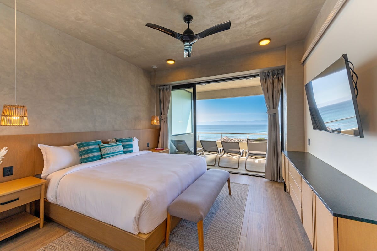 Una habitación de hotel moderna con una cama grande, un ventilador de techo, un televisor y un balcón con vista al océano.