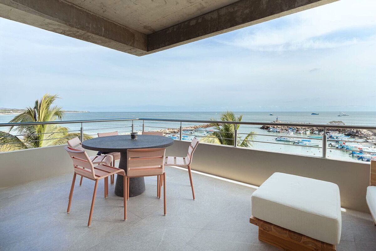 Un balcón con una mesa redonda y sillas, con vista a una playa con barcos y sombrillas.