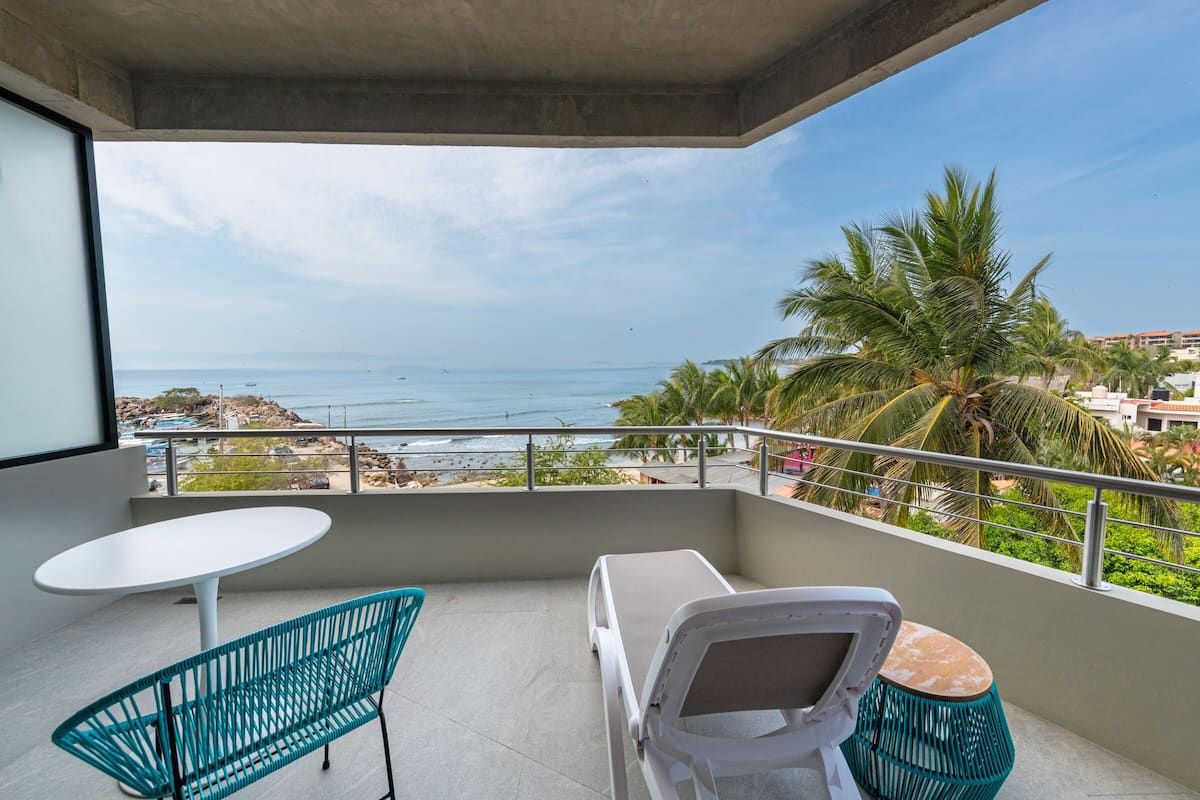 Un balcón con mesa blanca, una silla verde y una tumbona con vistas a la playa y al océano.