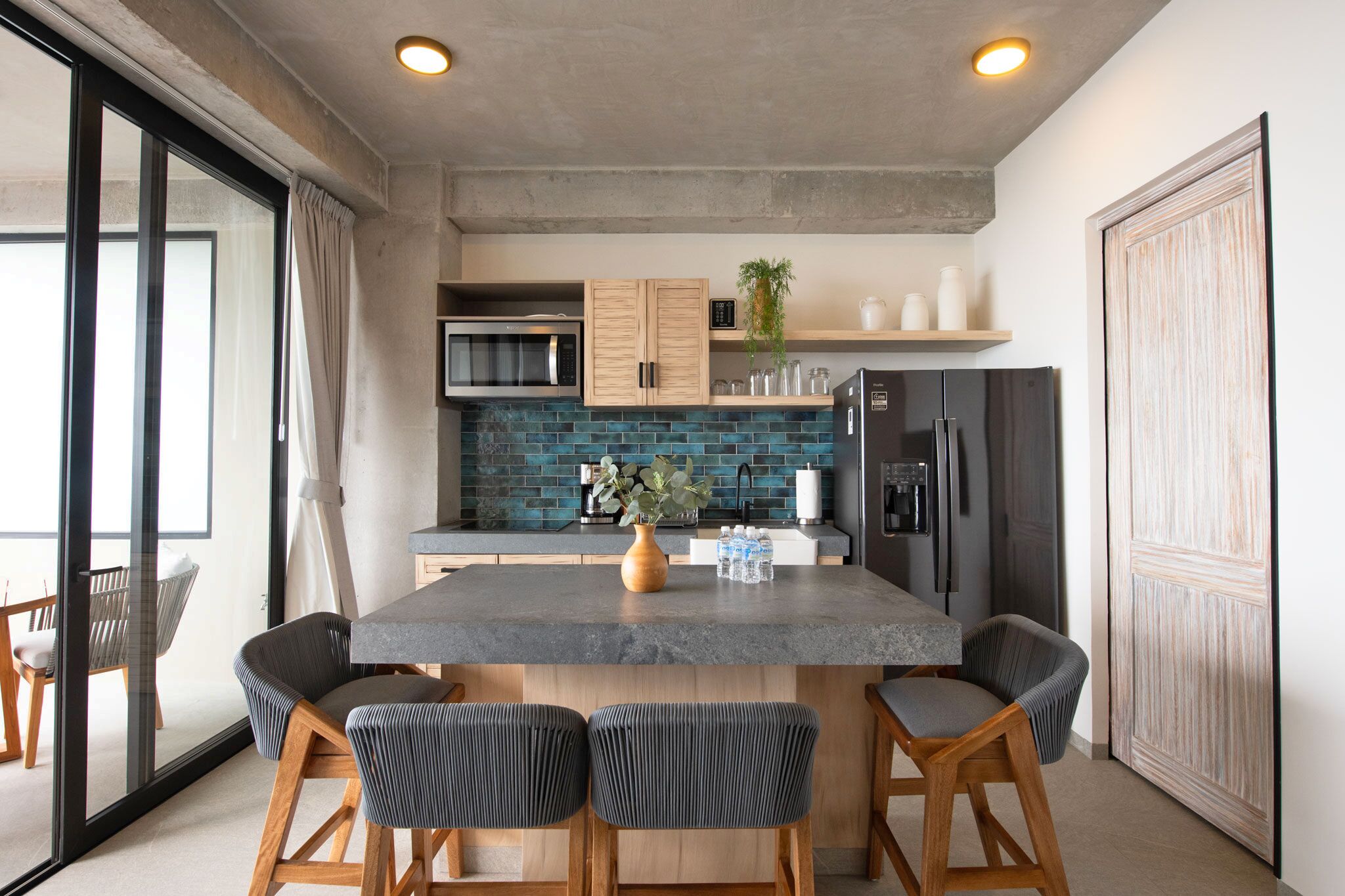A modern kitchen with a central island, wooden chairs, a microwave, a refrigerator, and a blue-tiled backsplash.