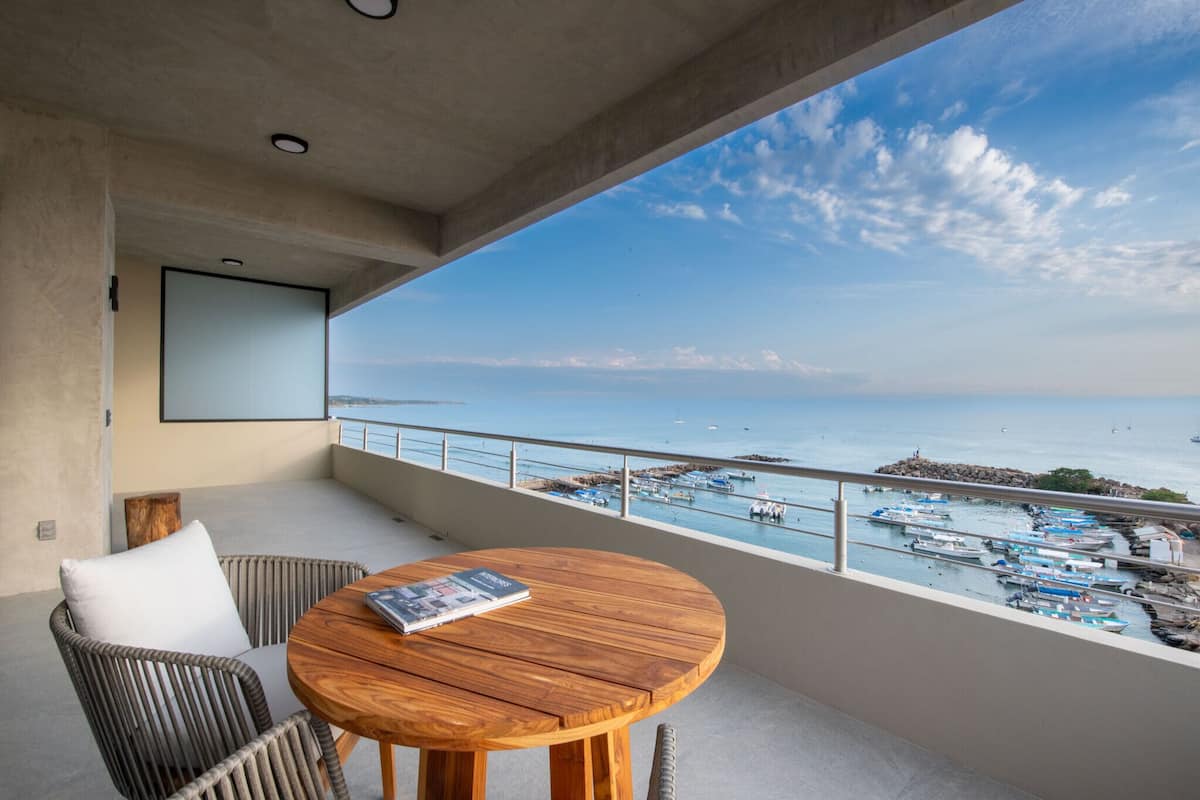 Un balcón con mesa de madera y sillas de mimbre, con vista a un puerto deportivo con barcos y cielo despejado.