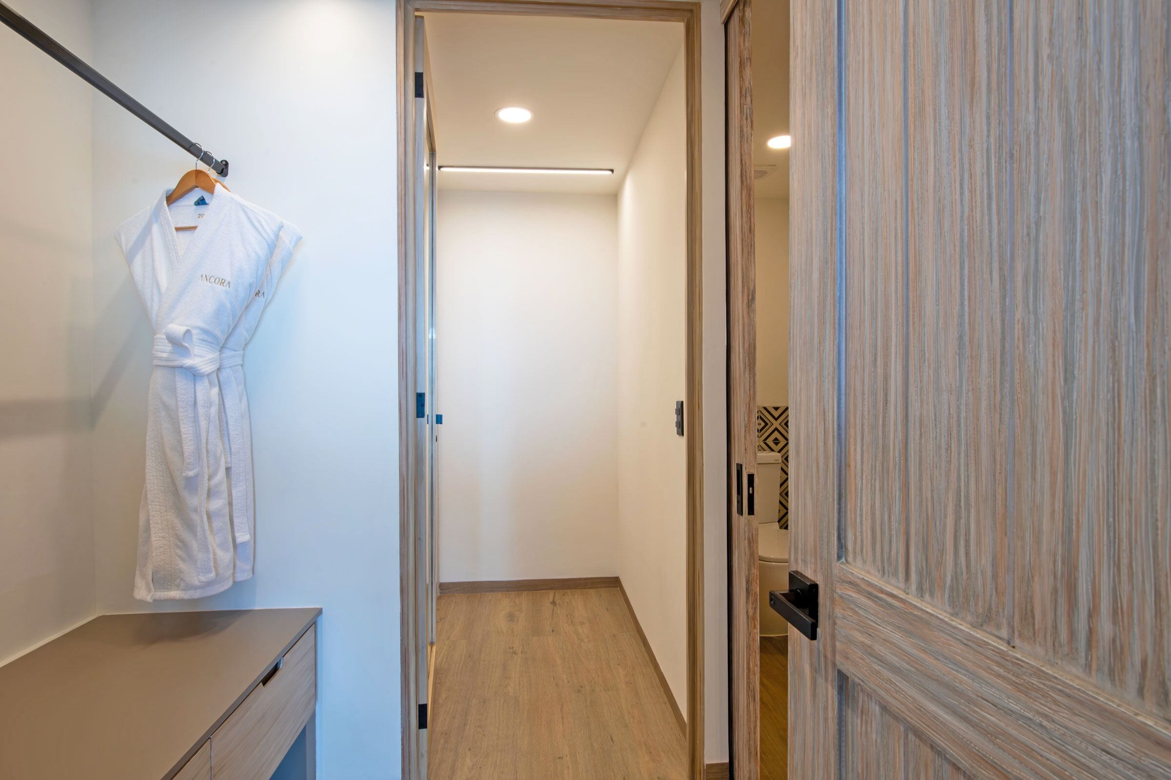 A hallway with a wooden floor, a white robe hanging on a rack, and a door with a patterned panel.