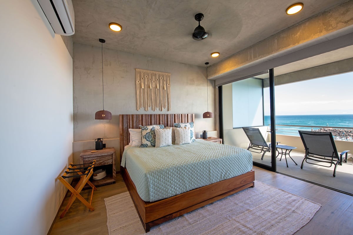 Un dormitorio con una cama grande, una mesita de noche, una silla de madera y vistas a la playa a través de amplias ventanas.