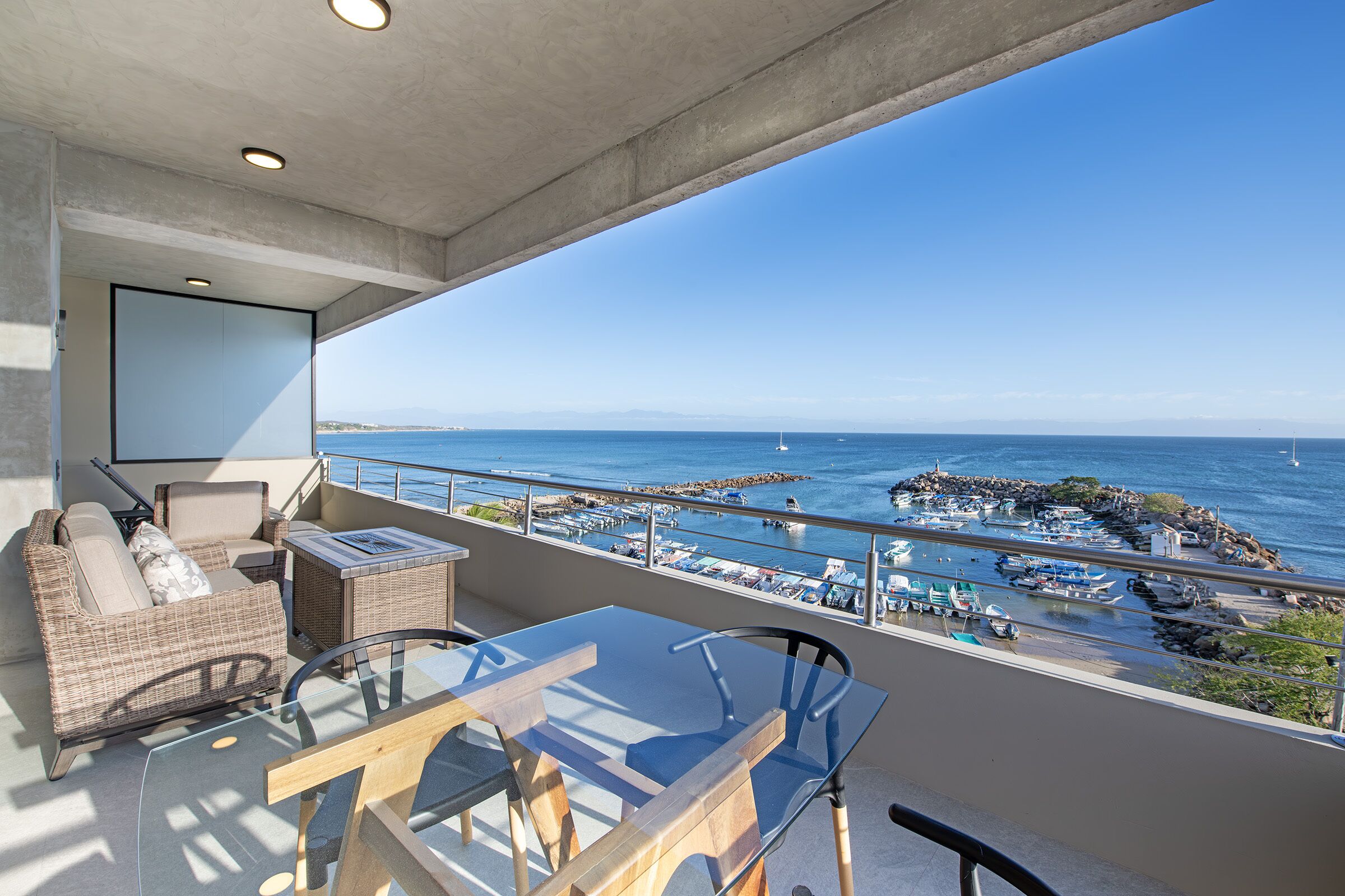 A balcony with a glass table, wicker furniture, and a view of the sea and boats.