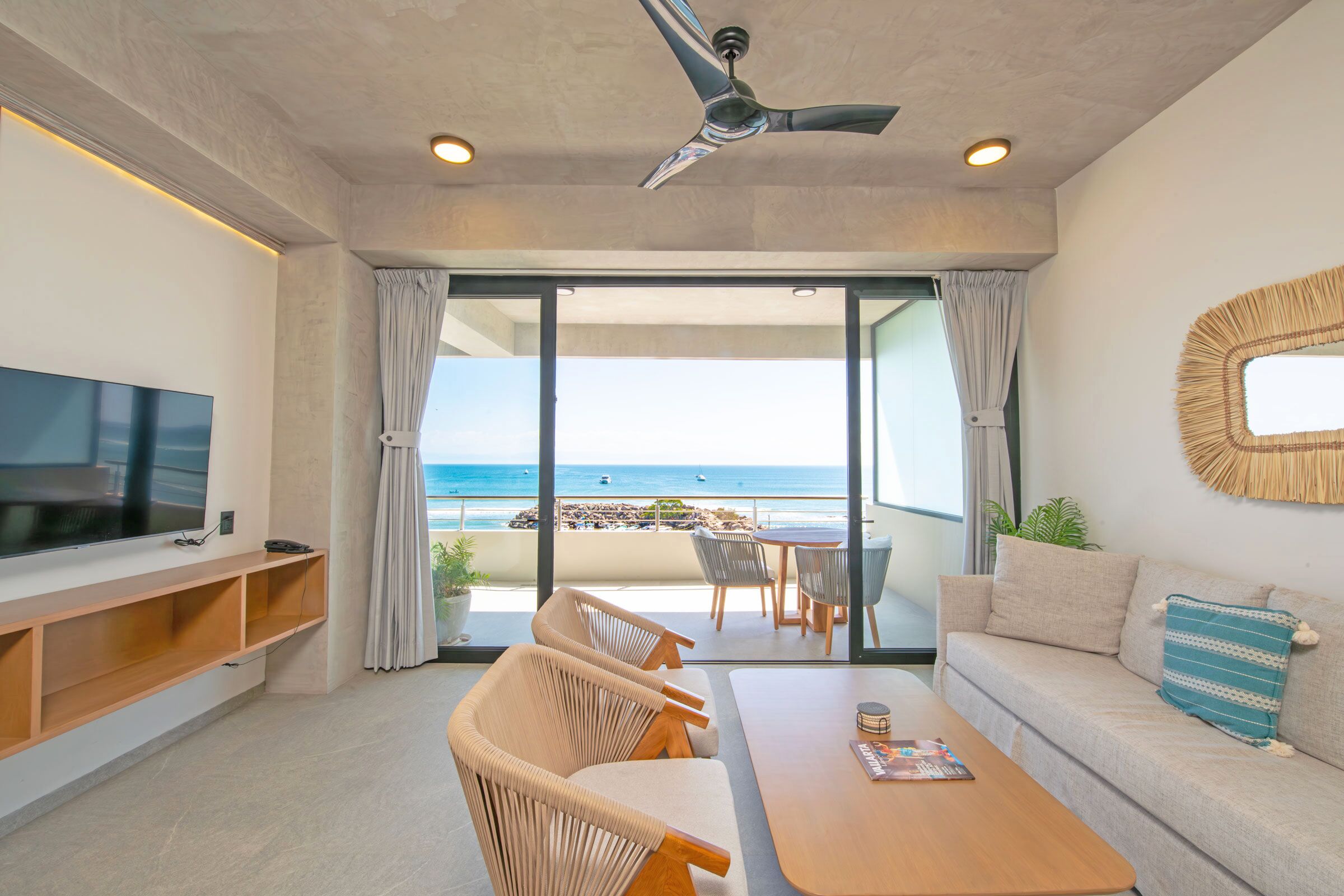 Una sala de estar moderna con televisor de pantalla plana, mueble de entretenimiento de madera, ventilador de techo, sofá con cojín decorativo estampado, mesa de centro y vista a la playa a través de grandes puertas corredizas de vidrio.