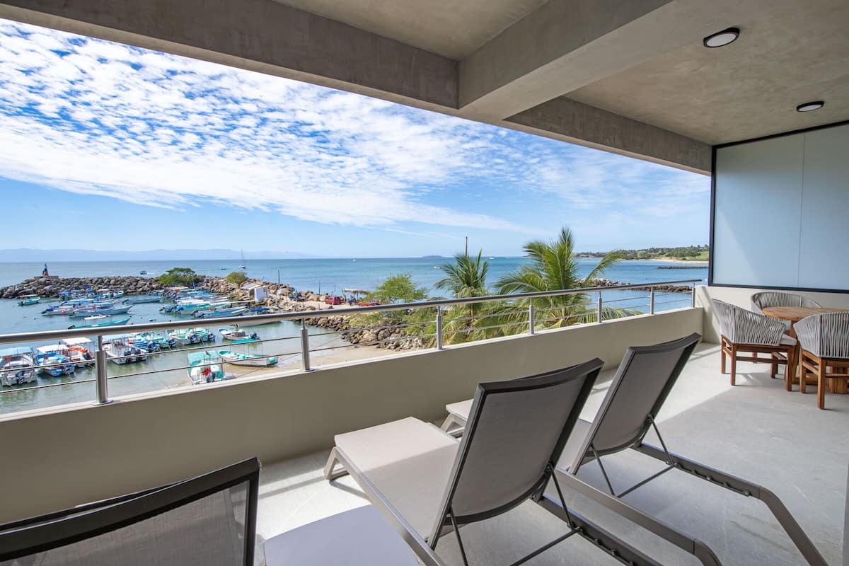 Un balcón con vista a barcos y a una playa, sillas de descanso y una mesa.