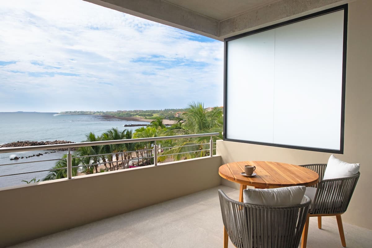 Un balcón con una mesa de madera y sillas de mimbre, con vistas a la costa, palmeras y una playa.