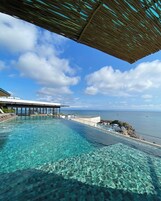 Una piscina infinita transparente con vista al océano y una zona cubierta.