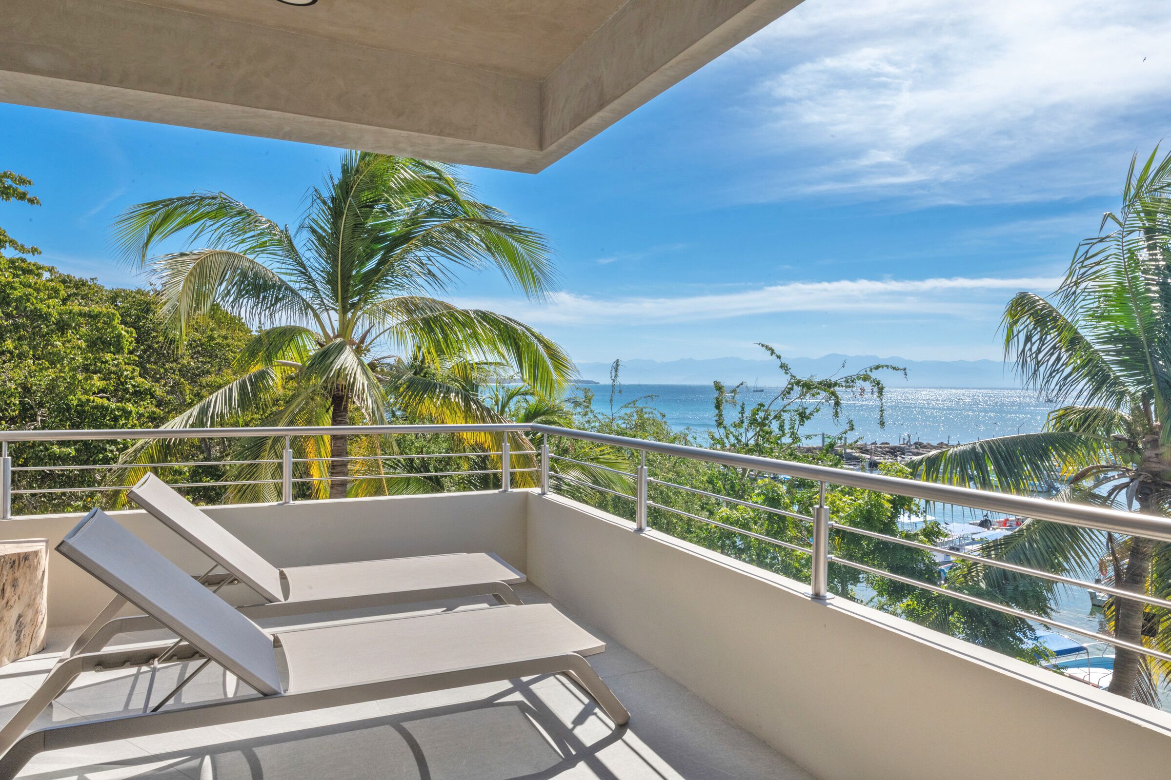 A balcony with two lounge chairs, a view of the sea, and palm trees.