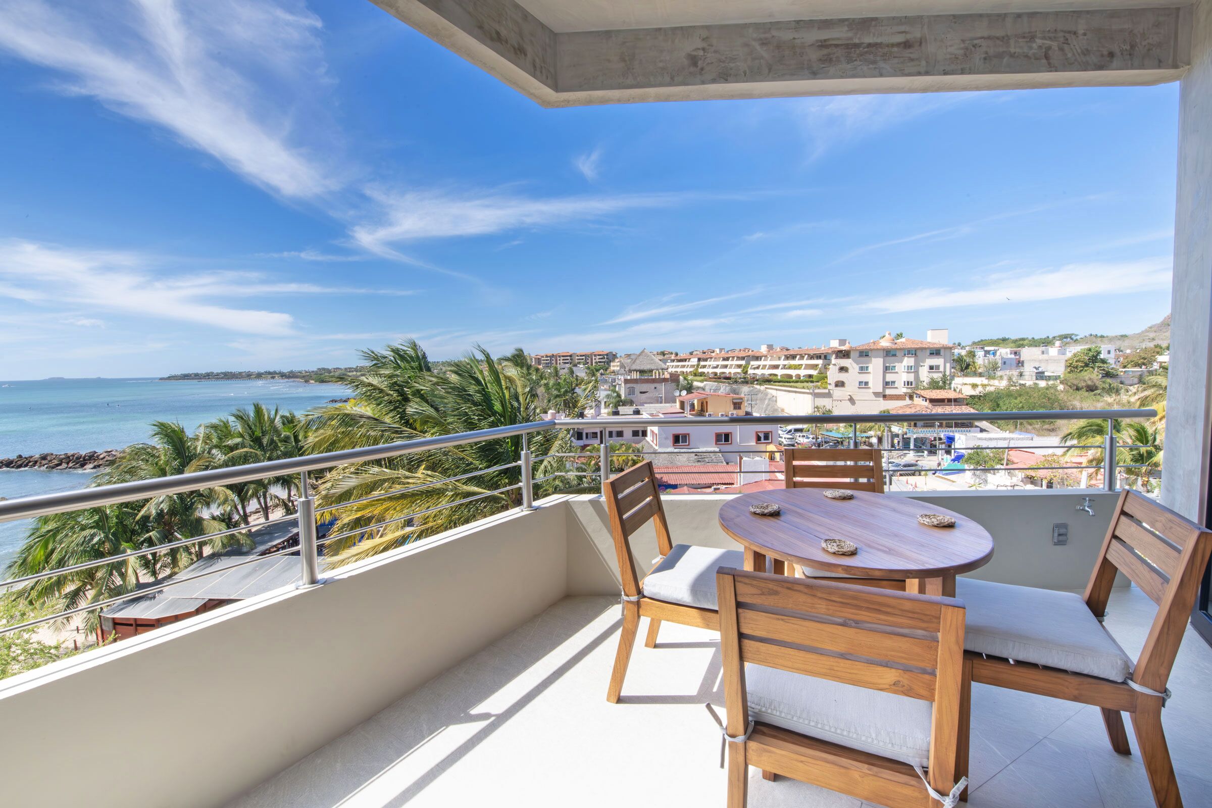 A balcony with a wooden table and chairs, overlooking a beach and a coastal town.