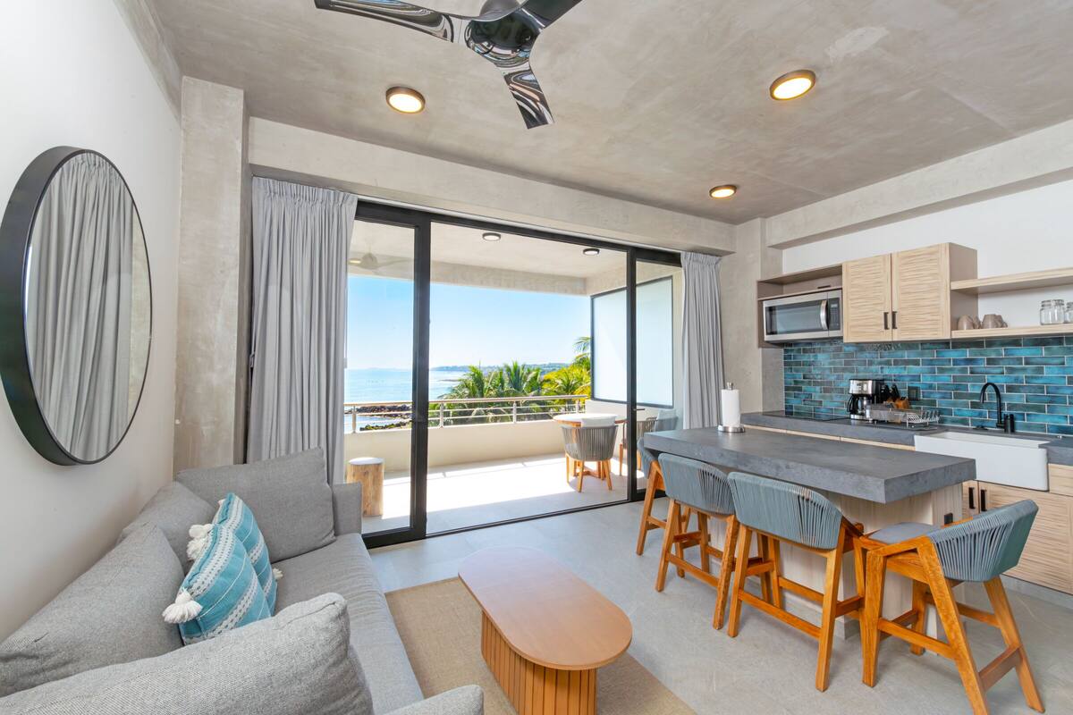 Una cocina moderna con una isla grande, un comedor con taburetes de bar, un sofá con cojines y vista al océano a través de puertas corredizas de vidrio.