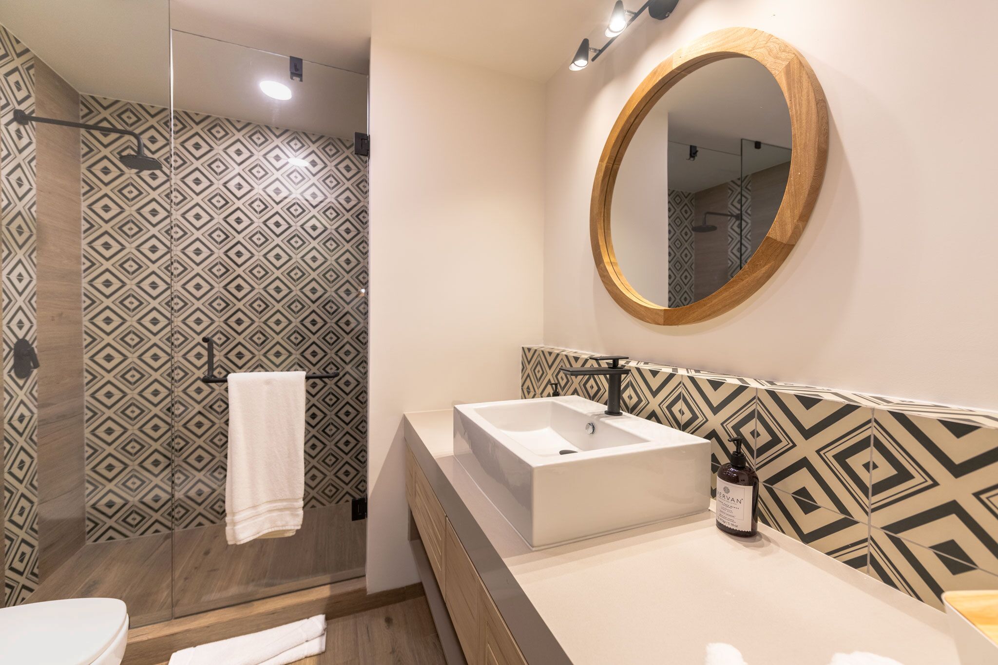 Un baño moderno con pared de diseño geométrico, un espejo con marco de madera, un lavamanos blanco y una toalla colgada en un perchero.