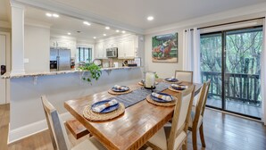 Un comedor con una mesa de madera dispuesta para cuatro, una cocina al fondo y un ventanal con vistas a árboles.