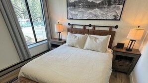 A bedroom with a large bed, two bedside tables with lamps, a bench, a patterned rug, and a framed picture on the wall. - Fireside Condo at the Village - Sleeps 4 - Newly Remodeled (Mammoth Lakes)
