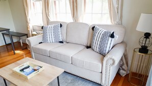 Una sala de estar con un sofá color beige, dos almohadones estampados, una mesita de centro de madera, una mesita auxiliar con una lámpara y una ventana con cortinas.